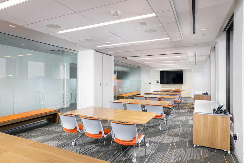 A modern conference room with rows of tables, orange-seated chairs, a wall-mounted flat-screen television, and glass walls.