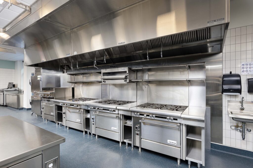 A row of commercial stainless steel gas ranges and ovens beneath a large ventilation hood in a kitchen.
