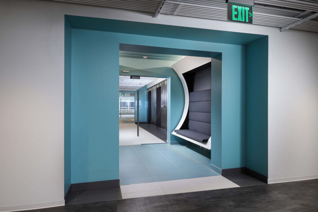 An office hallway with teal walls, a recessed gray cushioned bench, and an illuminated exit sign above the doorway.