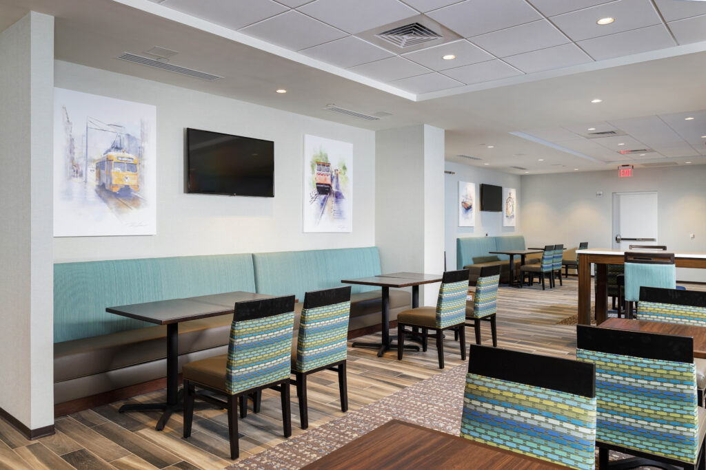 A hotel dining area with rows of turquoise booths, dark wood tables, and patterned chairs, decorated with framed paintings of cable cars.