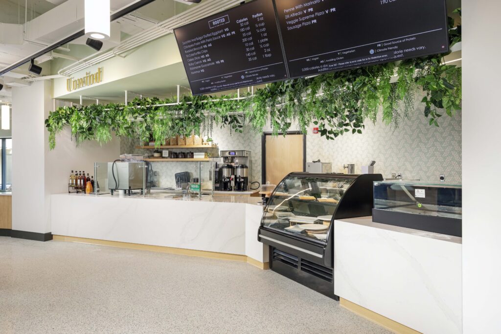 A cafe counter featuring a white marble top, hanging greenery, coffee machines, a glass display case, and a large digital menu board listing food items and nutritional information.