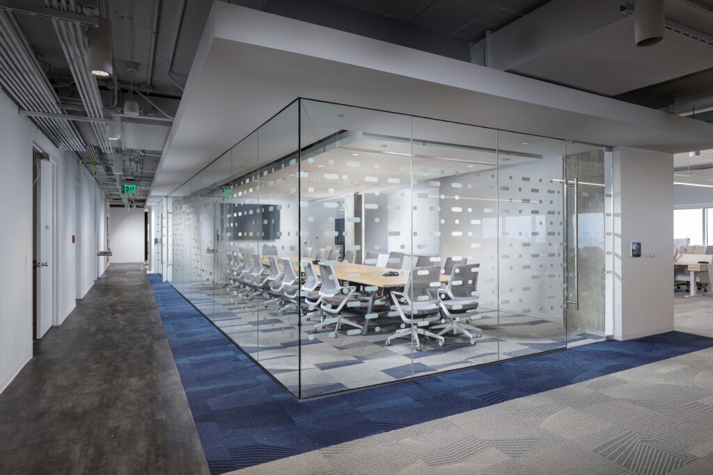 A glass-walled conference room with a long table and office chairs, situated in an open-plan office.