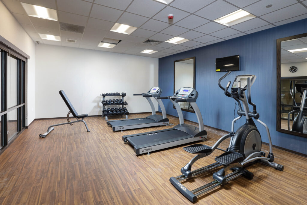 A gym interior with an adjustable weight bench, a rack of dumbbells, two treadmills, and an elliptical machine.