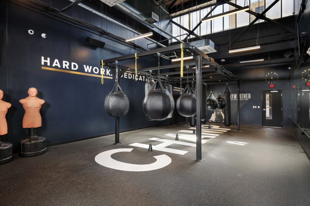Boxing gym interior with a central rack of hanging heavy bags, two freestanding punch dummies, and text on the wall reading “HARD WORK. DEDICATION.”