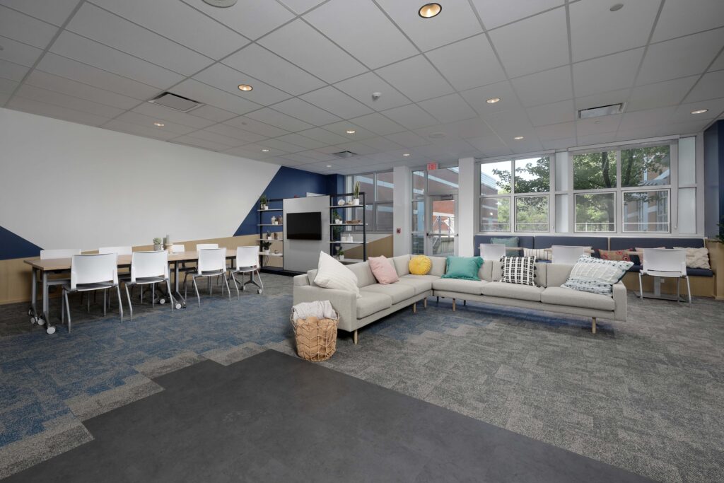 Modern office lounge area with a sectional sofa, a long conference table with chairs, and wall-mounted shelving.