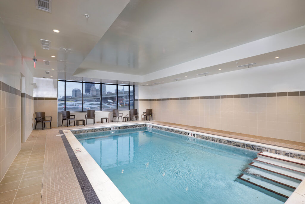 Indoor swimming pool with steps, surrounded by beige tile walls and chairs facing a window with a city view.