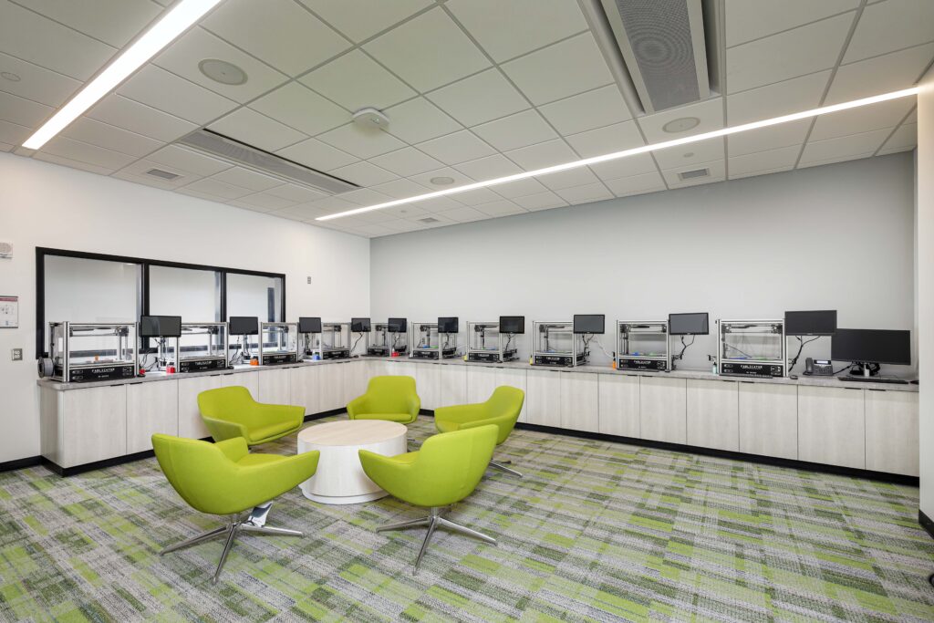 A room with a row of 3D printers on a counter and a seating area with four lime green chairs around a circular table.