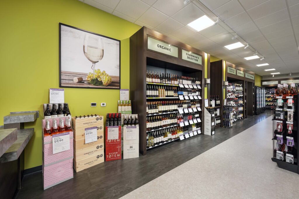 Wine shop aisle with shelves of bottles labeled “Organic” and “Chairman’s Selection,” featuring floor displays of assorted wines against a bright green wall.