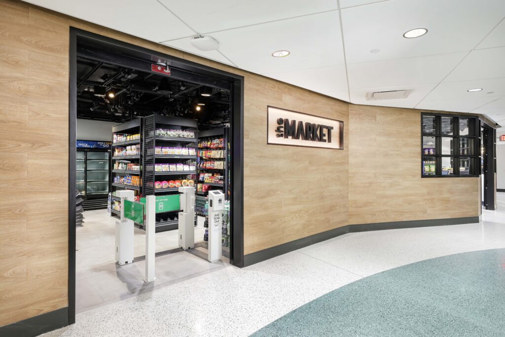 Entrance to a “The Market” convenience store with automated gates and shelves stocked with snacks and drinks.