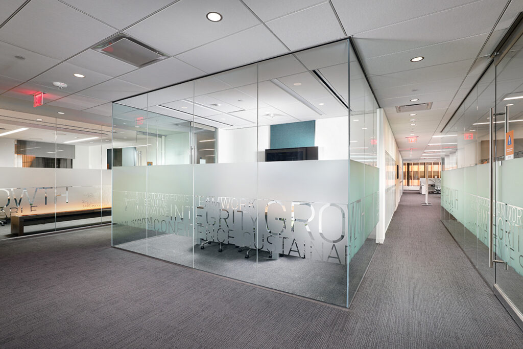 A glass-walled office conference room in an open-plan workspace, with words like “Growth,” “Integrity,” and “Teamwork” etched into the frosted glass panels.