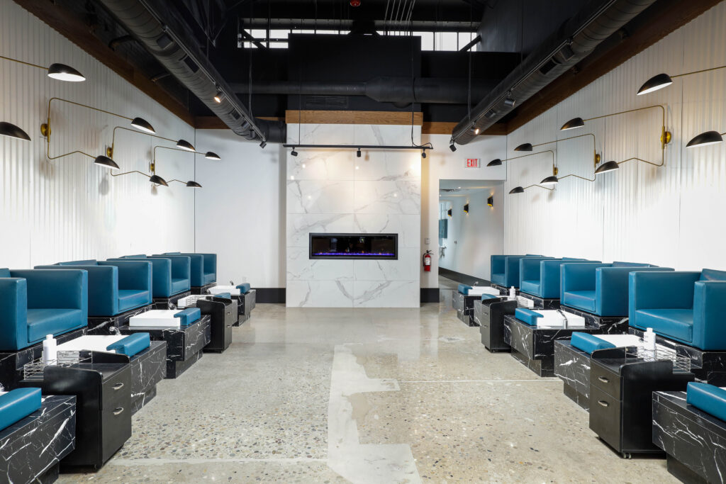 A nail salon interior with two rows of teal pedicure chairs on black marble bases facing a central white marble fireplace.
