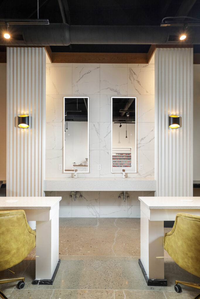 Two manicure stations with yellow chairs facing a marble-tiled wall featuring two rectangular mirrors, wall sconces, and a nail polish rack.
