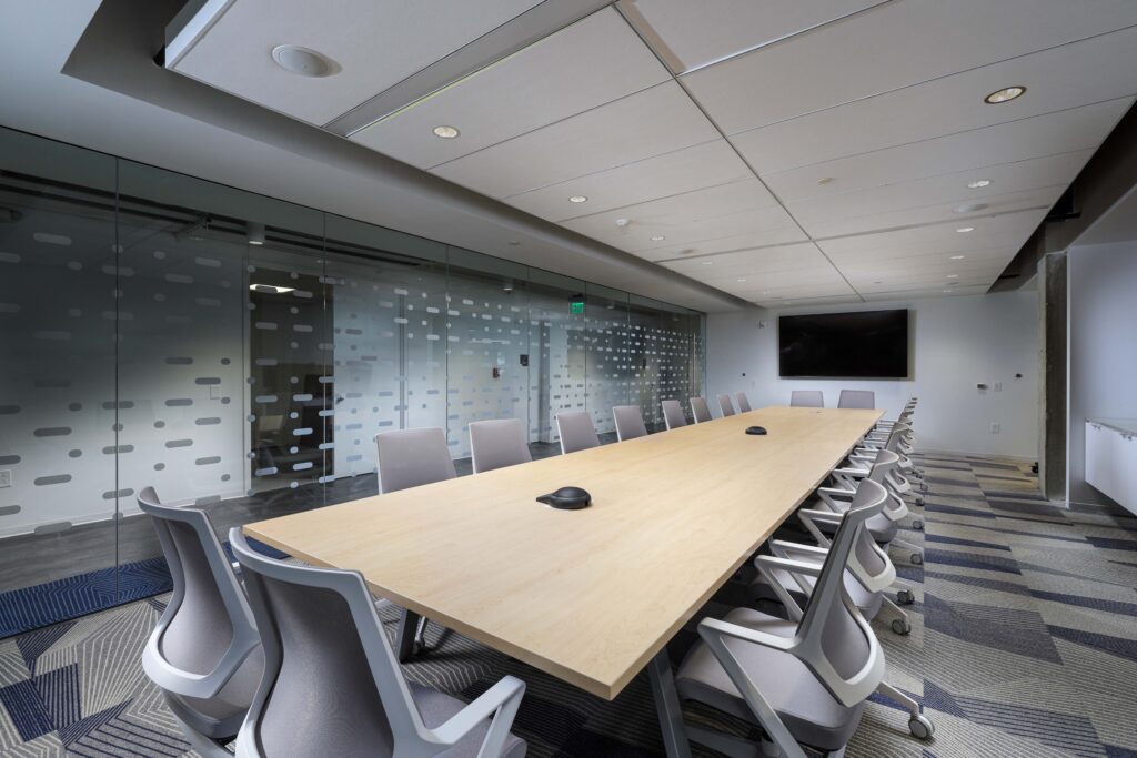 A long light-wood conference table with grey office chairs in a modern meeting room with glass walls.