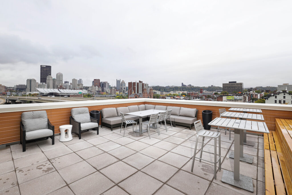Rooftop patio with outdoor seating, tables, and a view of the Pittsburgh skyline under a cloudy sky.