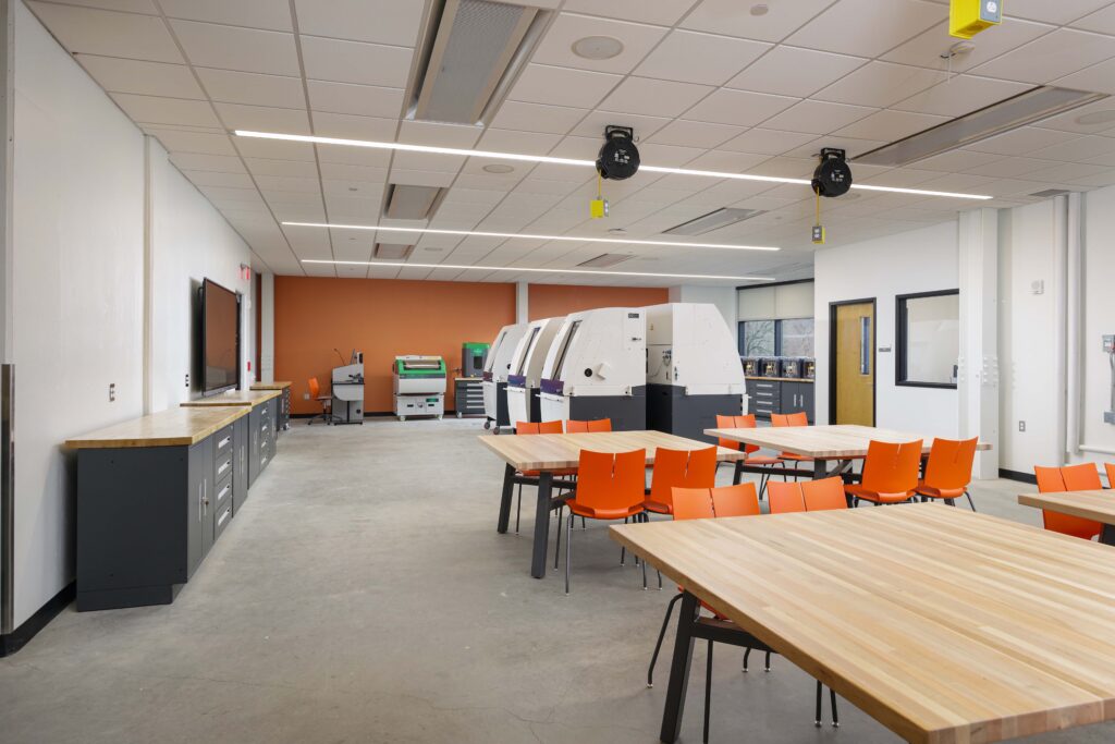 A fabrication lab with wooden tables, orange chairs, and several large pieces of industrial machinery in the background.
