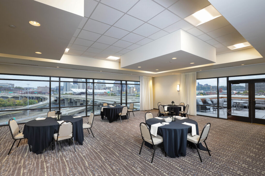 A banquet room with round tables covered in black tablecloths and chairs, featuring large windows overlooking a city highway and skyline.
