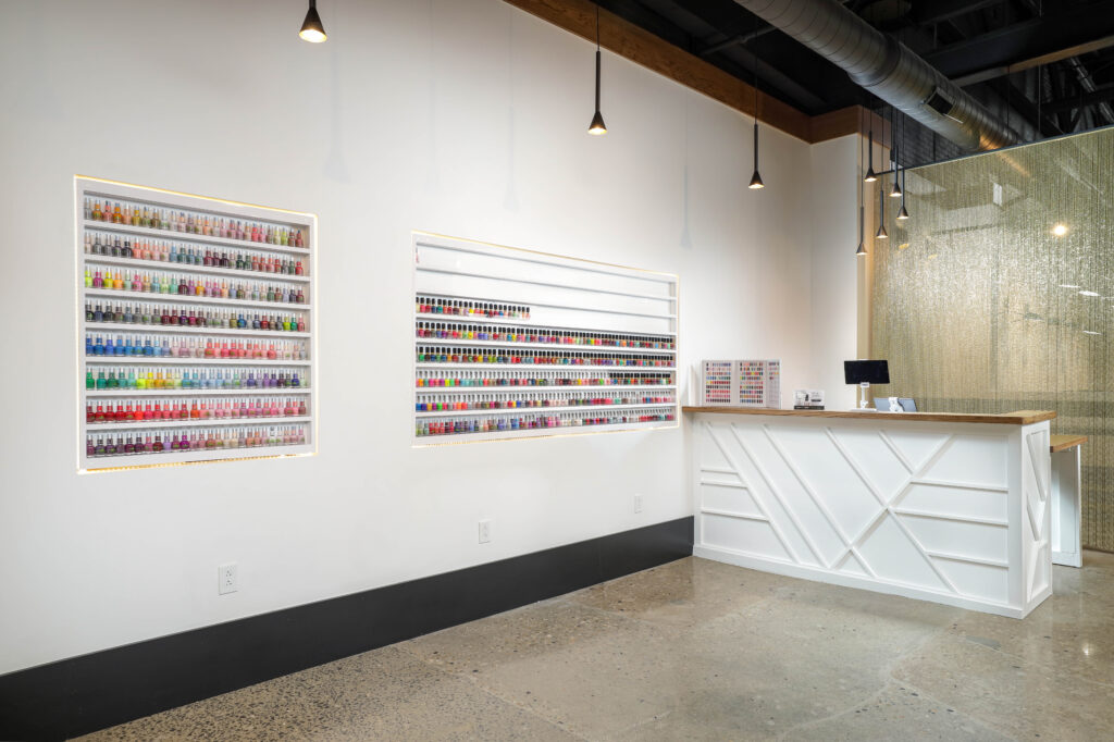 A nail salon reception area featuring wall-mounted shelves displaying rows of colorful nail polish bottles next to a white service desk.