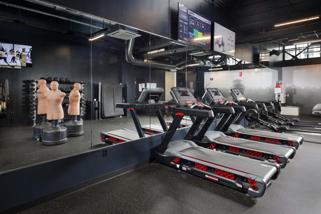 Row of treadmills in a gym with two grappling dummies in the background and a television screen displaying workout metrics.