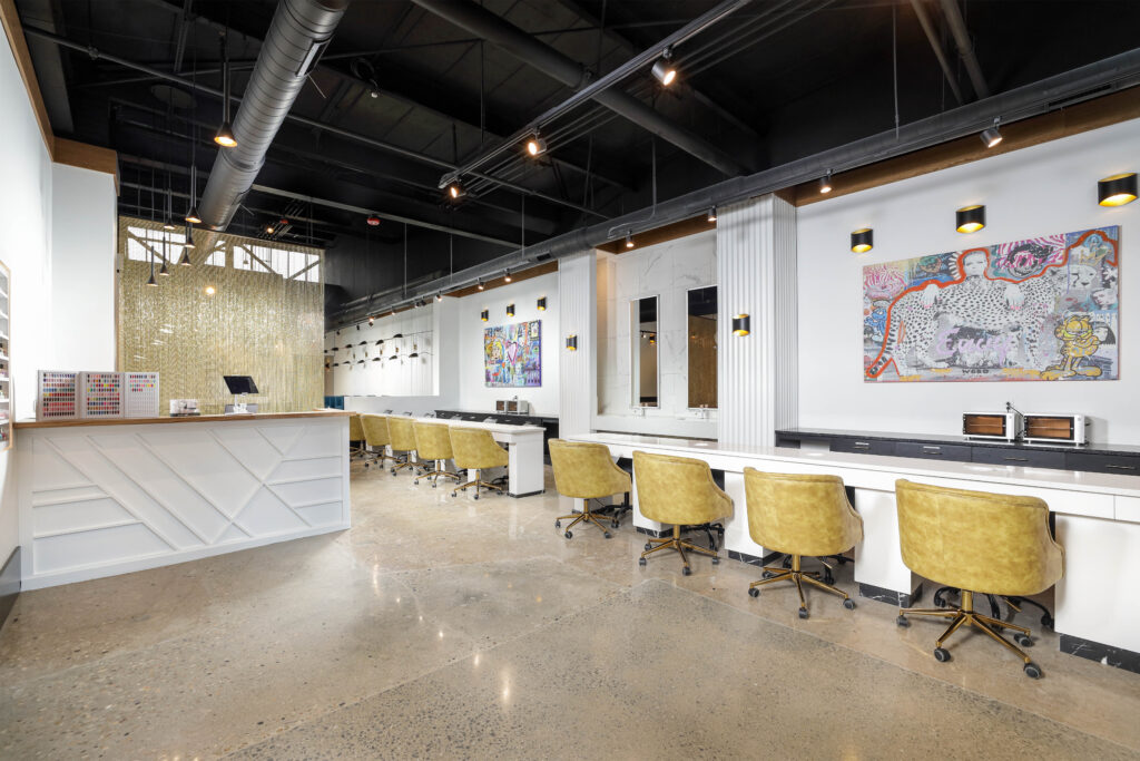 A modern hair salon with a white reception desk, a long row of workstations with tan chairs, and colorful artwork on the walls.