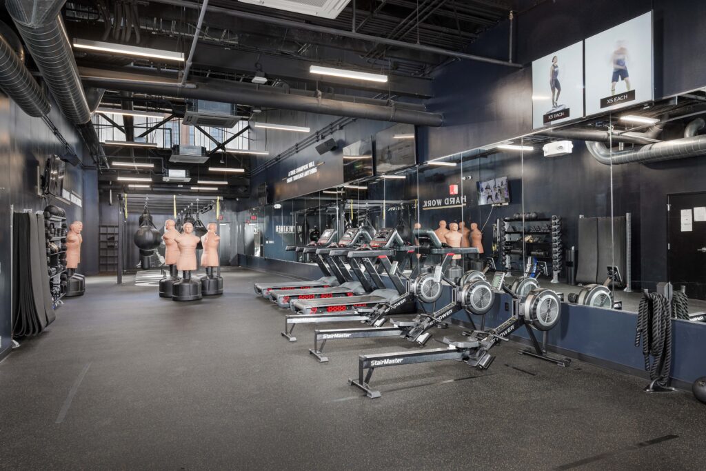 Gym interior with rowing machines, treadmills, and freestanding punching dummies reflected in a large wall mirror.