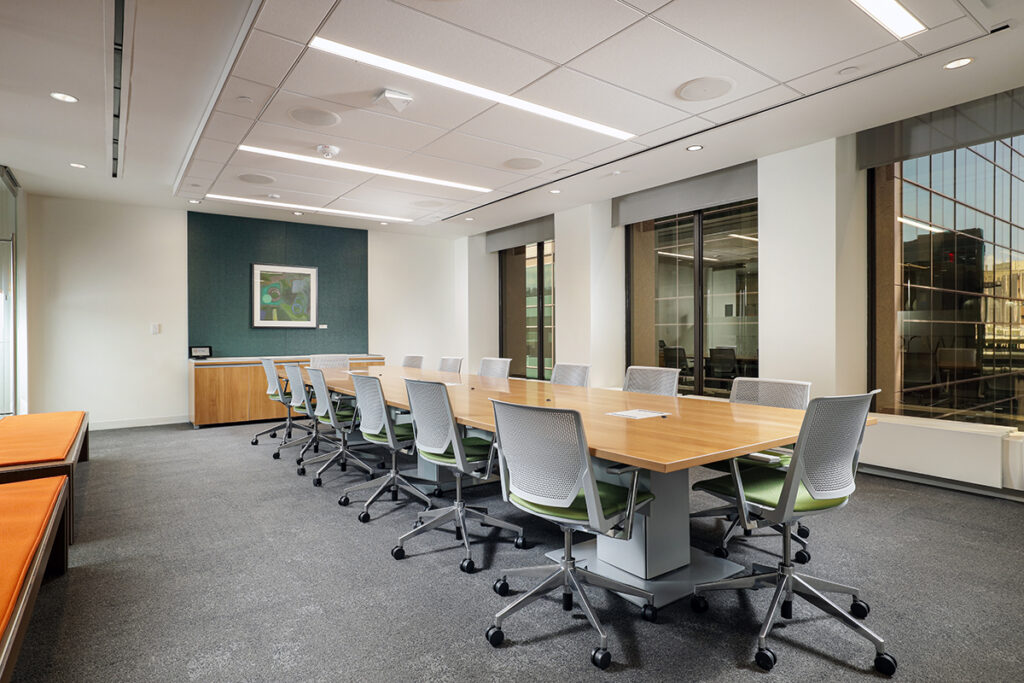 A modern conference room with a long wood table surrounded by ergonomic rolling chairs and a framed abstract painting on the back wall.