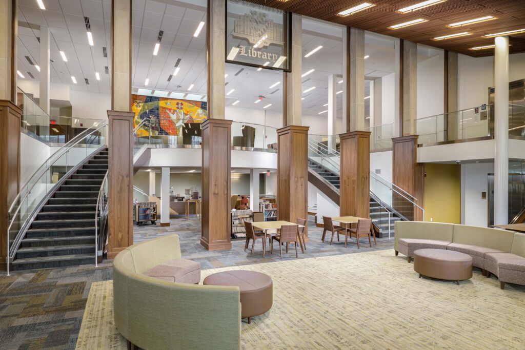 A bright library lobby with two symmetrical staircases, seating areas featuring curved sofas and small tables, and a large colorful mural on the upper level.