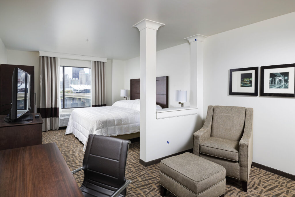 Hotel room with a bed, a desk chair, an armchair with an ottoman, and a window view of a city skyline.