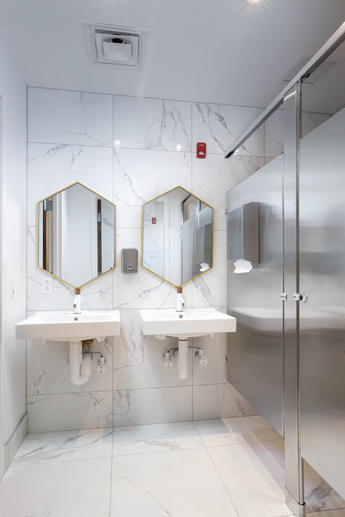 Two white wall-mounted sinks with hexagonal gold-rimmed mirrors in a bathroom with marble-tiled walls.