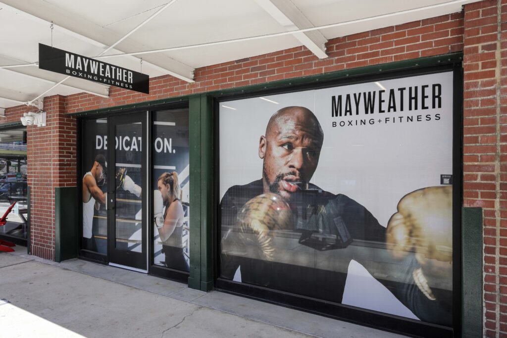 Storefront of a Mayweather Boxing + Fitness gym with large window decals featuring images of boxers and the company name.