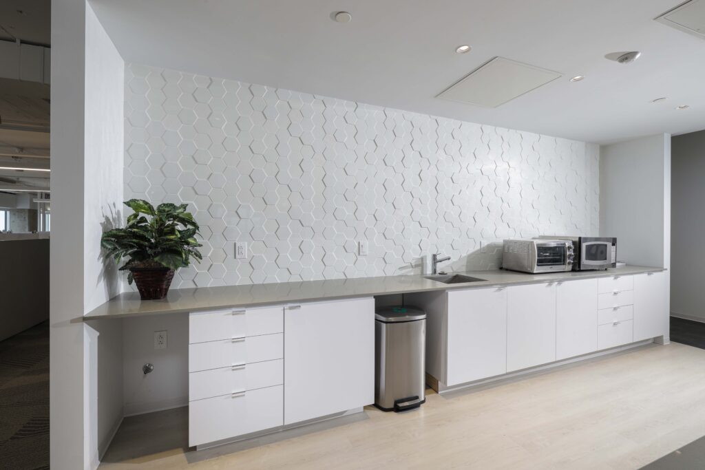 Office kitchenette with white cabinets, light countertops, a stainless steel trash can, a toaster oven, a microwave, and a white hexagonal tile backsplash.