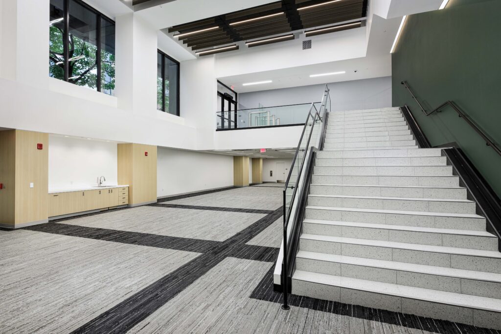 A wide, modern interior lobby featuring a large staircase with white treads and a glass railing, leading to a second-floor landing.