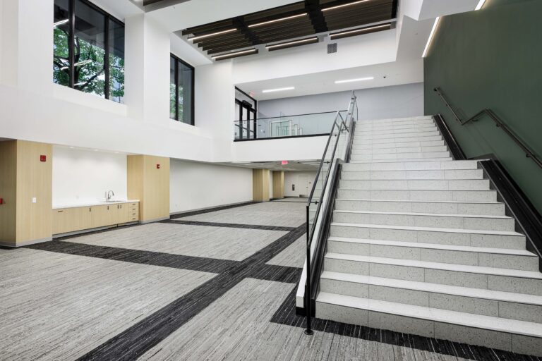 A wide, modern interior lobby featuring a large staircase with white treads and a glass railing, leading to a second-floor landing.
