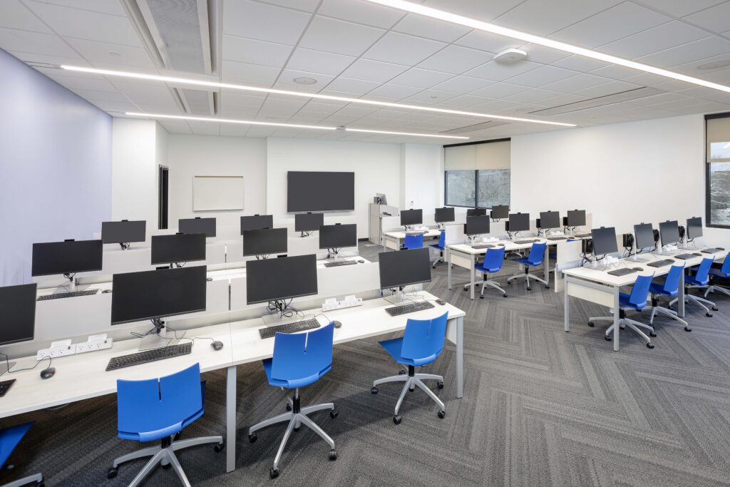 A computer lab with rows of desks, each equipped with a monitor, keyboard, and blue rolling chair.