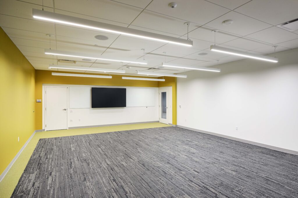 An empty meeting room with gray carpet, white walls, a yellow accent wall, and a large wall-mounted monitor.