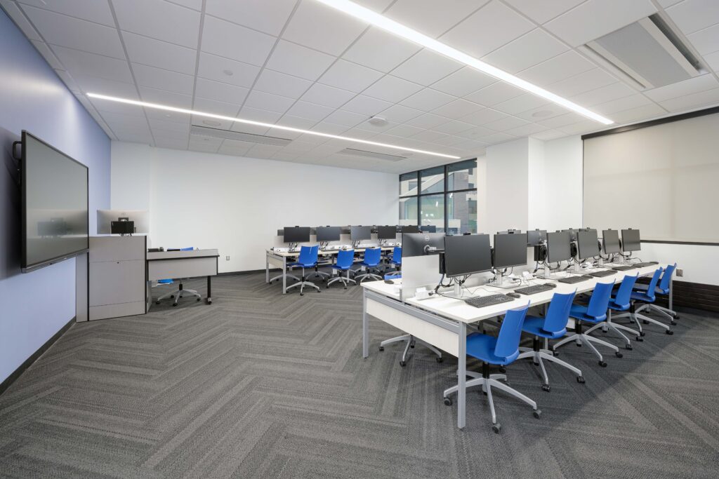 A modern computer lab with rows of desks, blue chairs, and desktop computers facing a wall-mounted display.