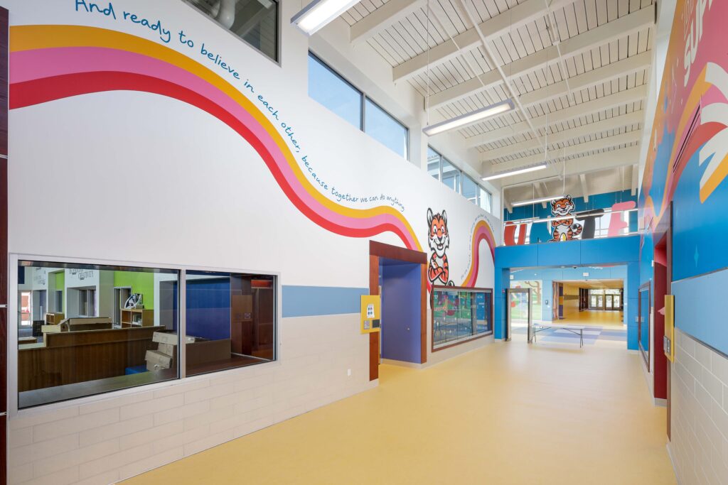 School hallway with yellow floors, featuring a wall mural of colorful swooping lines, a cartoon tiger mascot, and the text “And ready to believe in each other, because together we can do anything.”