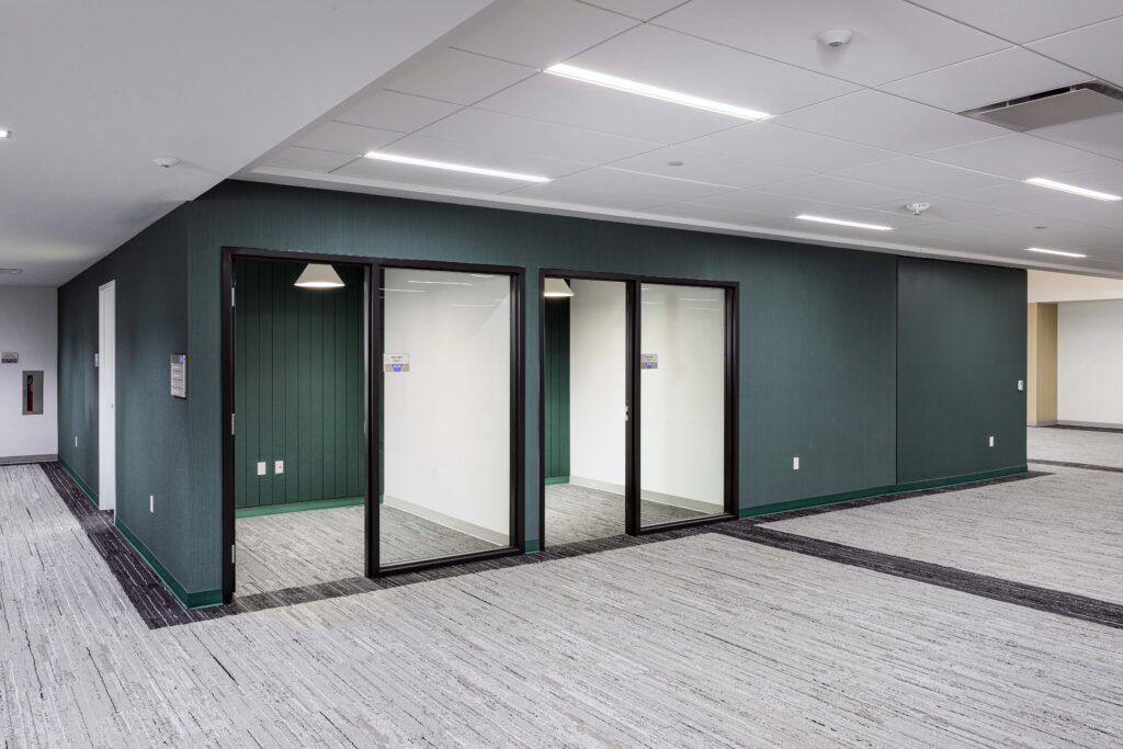 Office interior featuring two glass-walled meeting rooms against a dark teal wall and gray patterned carpet.