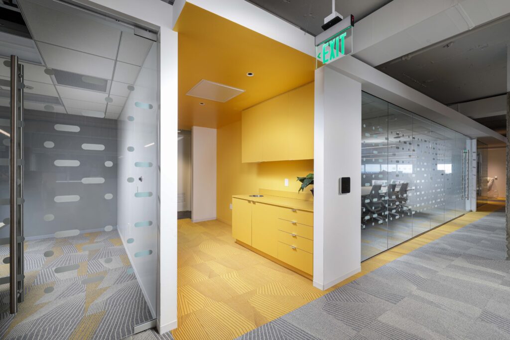 An office kitchenette with yellow cabinets and flooring, flanked by glass-walled conference rooms.