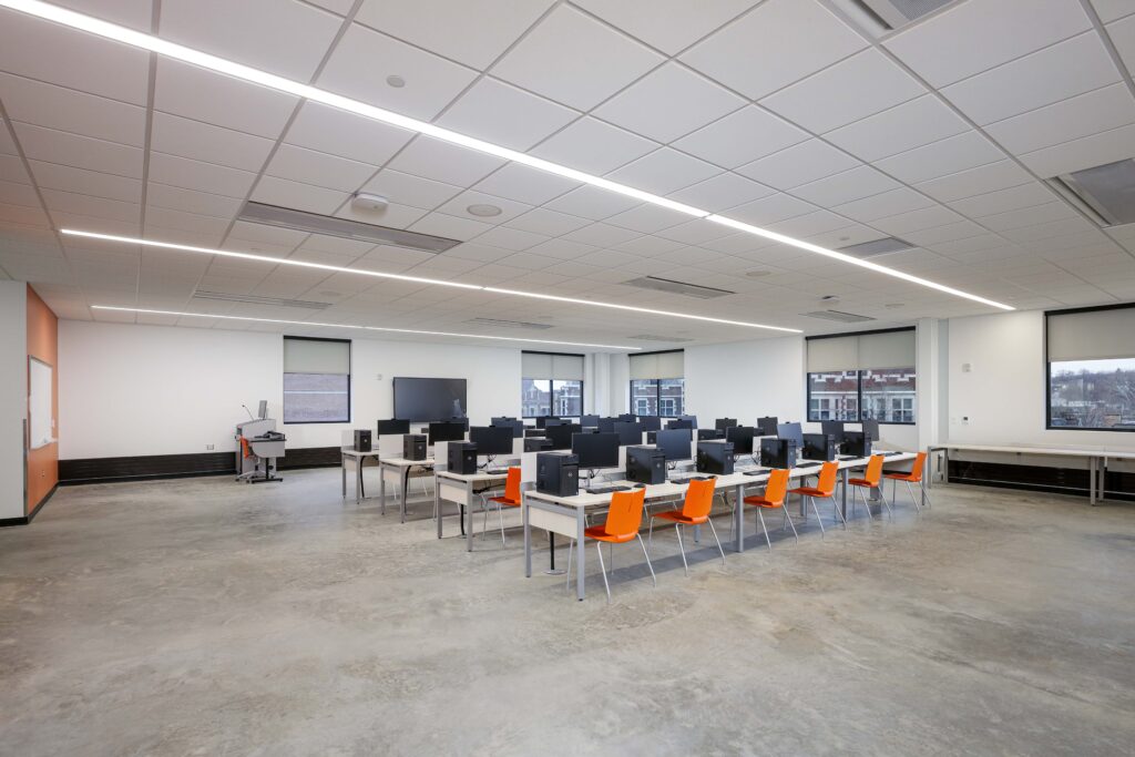 A computer lab with rows of desks, black monitors, and orange chairs on a concrete floor.
