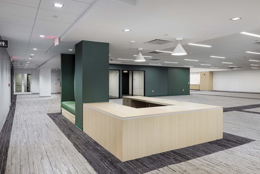 A modern office reception area with a light wood L-shaped desk, a dark green wall, and a built-in green bench.