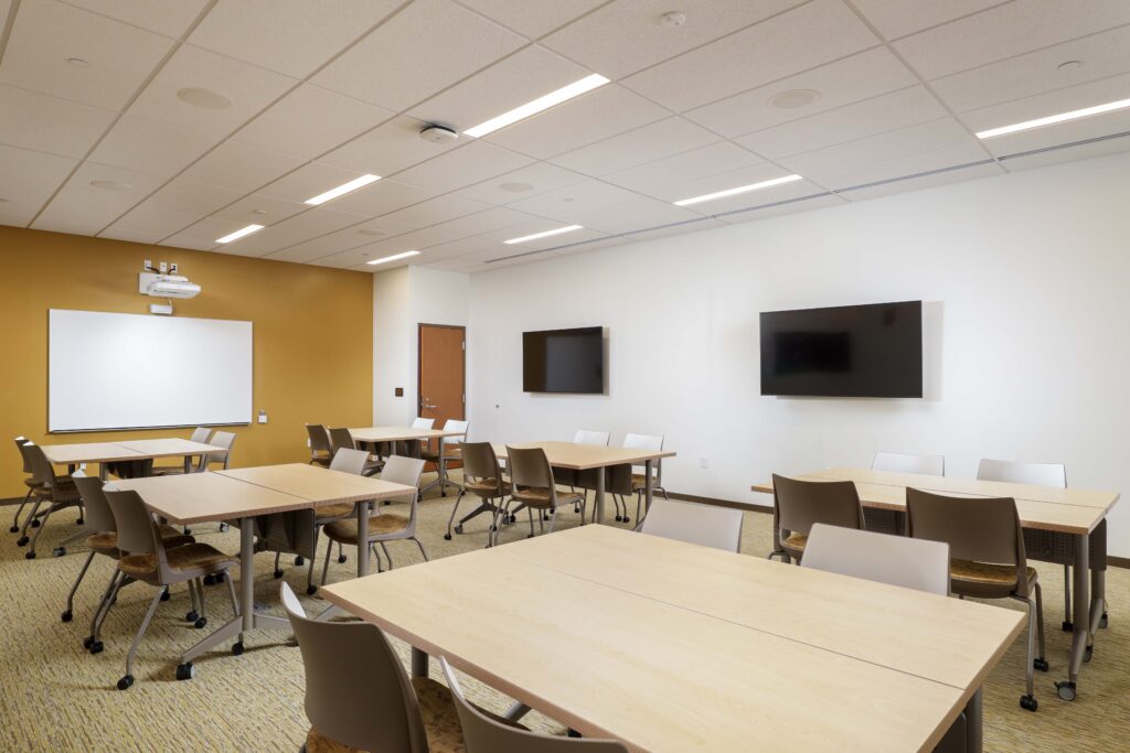 A classroom with multiple light-wood tables, gray chairs, a whiteboard on a mustard wall, and two wall-mounted televisions on a white wall.