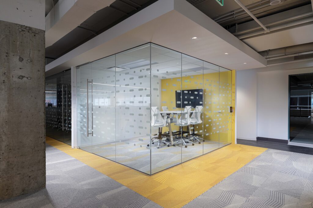 A glass-walled conference room with white chairs, a table, and a monitor, situated on a floor with yellow and gray patterned carpet.