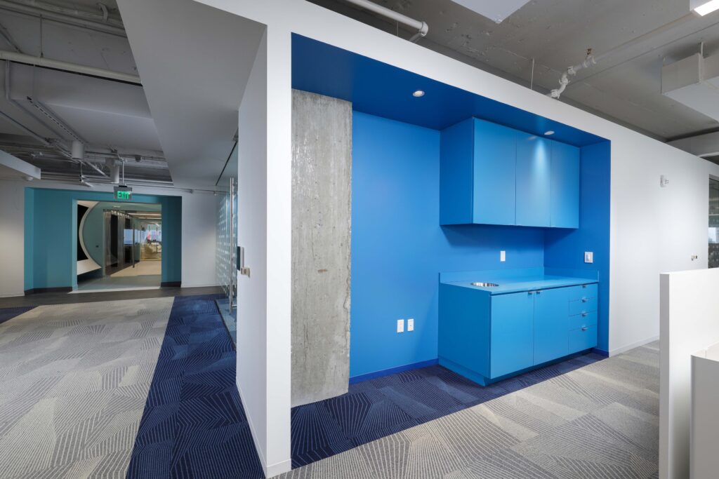 A blue office kitchenette with matching cabinets and a small sink, set against a bright blue accent wall and a concrete pillar.