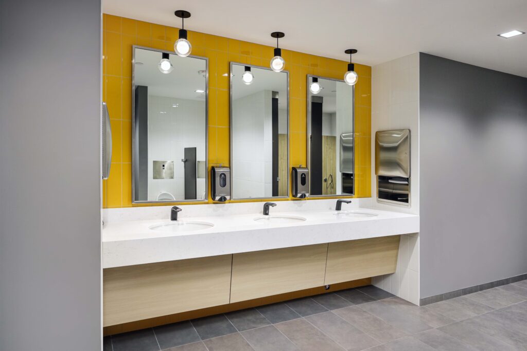 Three bathroom sinks with mirrors and pendant lights against a yellow tiled wall, with soap dispensers and a paper towel dispenser nearby.