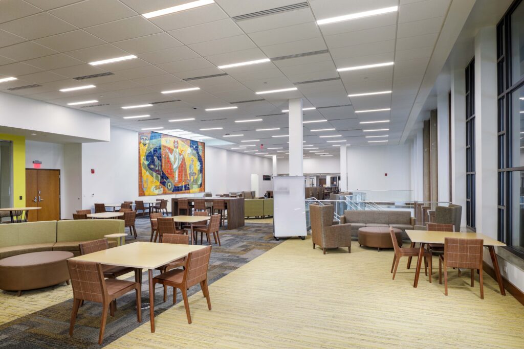 Modern lobby or lounge area with multiple tables, chairs, sofas, a whiteboard, and a large colorful mural on the far wall.