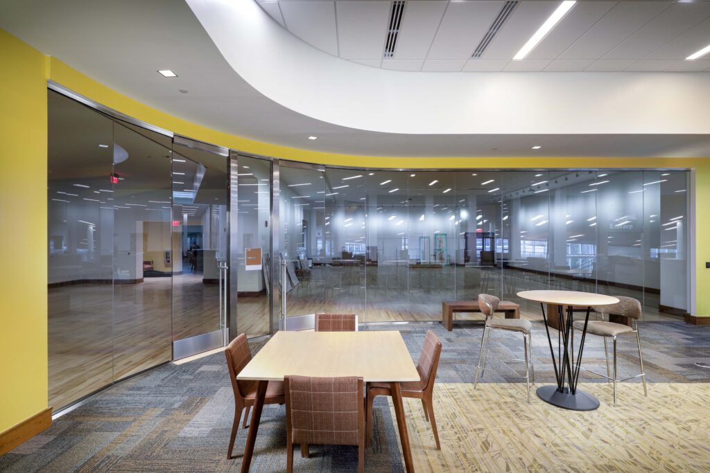 A modern interior lounge with a square dining table, a high bistro table with bar stools, and a glass-walled conference room.