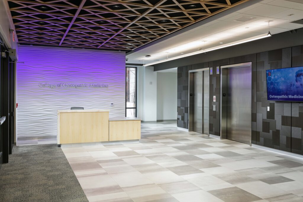 Reception area of the College of Osteopathic Medicine with a light wood desk, a textured white wall, two elevators, and a digital display screen.
