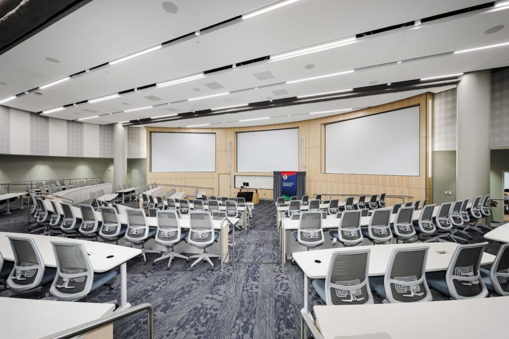 A large, tiered university lecture hall featuring rows of white desks and grey mesh chairs facing a stage with three projection screens and a College of Medicine lectern.