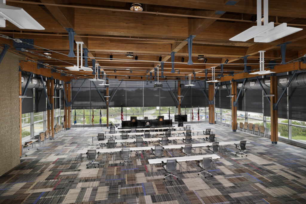 A large, open conference room with rows of tables and chairs facing a raised dais with flags in front of floor-to-ceiling windows.