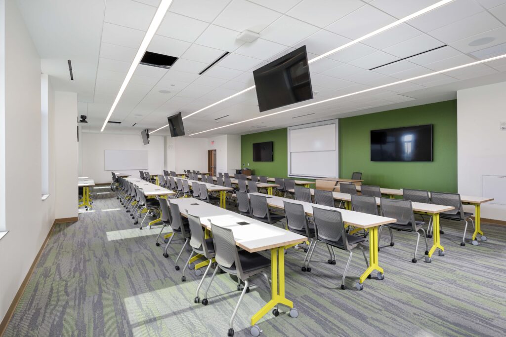 A classroom featuring rows of tables and chairs, wall-mounted screens, and whiteboards.